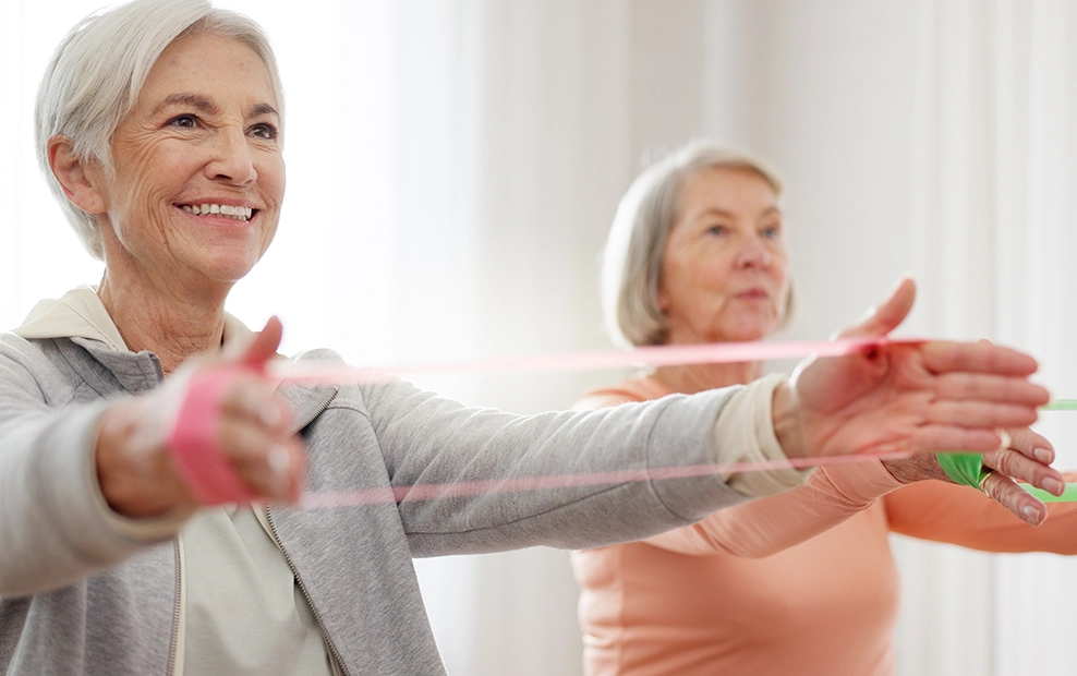 Two senior women performing resistance band exercises, demonstrating the effective therapy programs at Pivot Rehabilitation. The focus is on delivering high-quality, patient-centered care and improving patient outcomes through tailored physical therapy solutions.